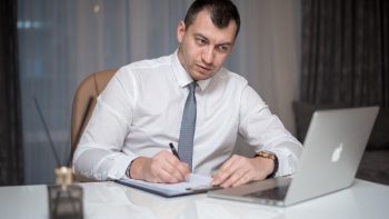 Focused businessman in white shirt writing notes at desk with laptop.