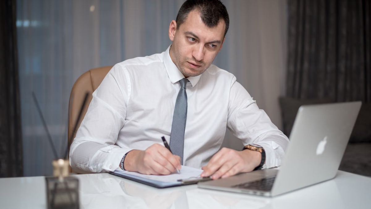 Focused businessman in white shirt writing notes at desk with laptop.